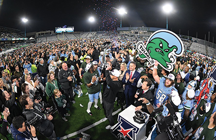 Fans celebrate on a football field at night, confetti falling, person holding a Tulane Angry Wave logo.