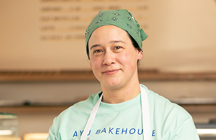 Smiling woman in a green bandana and "AYU BAKEHOUSE" apron, standing in a bakery.