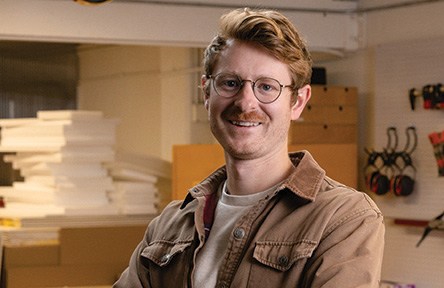 Smiling man with glasses and mustache in a workshop.