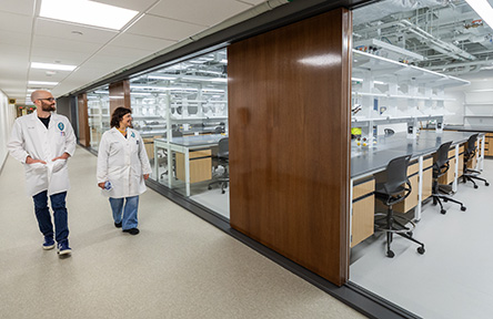 Two scientists in lab coats walk past a large, modern lab with glass walls.
