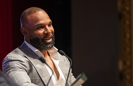 Man with beard and gray suit smiling, speaking into microphone at an event.