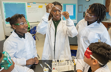 Four students in lab coats and goggles conduct a science experiment with a dropper.