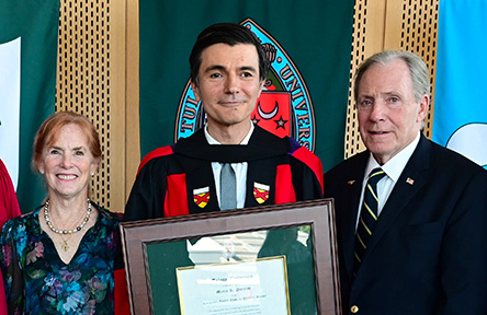 Man in academic regalia, flanked by two people, holds a framed certificate.
