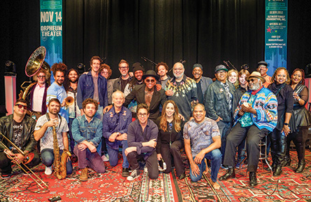 Large group of diverse musicians, some holding instruments, pose together on a stage.