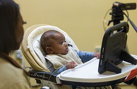 Baby in high chair watching a tablet, with an adult and camera nearby.