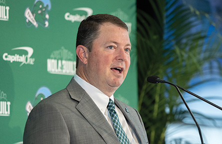 Coach in a suit speaks at a podium, green background with event logos.