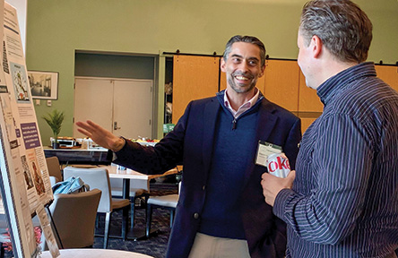 Smiling man in blue blazer explains poster to another man at an indoor event.