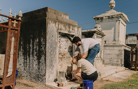 Man and woman working on an old, weathered tomb in a sunny cemetery.