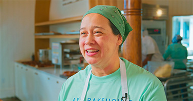 Happy woman in green bandana and apron smiles in a bakery.