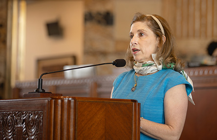 A woman in a blue dress speaks passionately at a podium with a microphone. She wears a scarf and headband, conveying a focused and engaged demeanor.