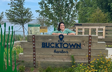 Person smiling behind a wooden sign that reads "Bucktown Harbor." Surrounding the sign are trees, greenery, yellow flowers, and a view of water.