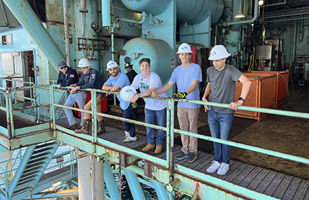 A group of six people in hard hats and casual clothes stands on a metal platform of an offshore industrial structure.