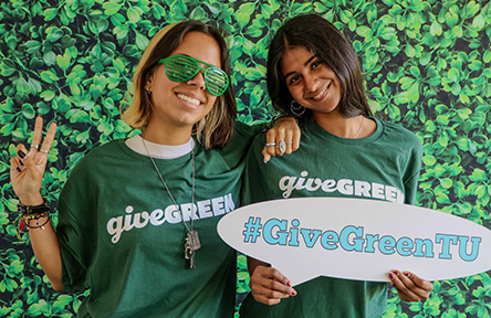 Two smiling women in "givegreen" shirts, one with sunglasses, holding a "#GiveGreenTU" sign.