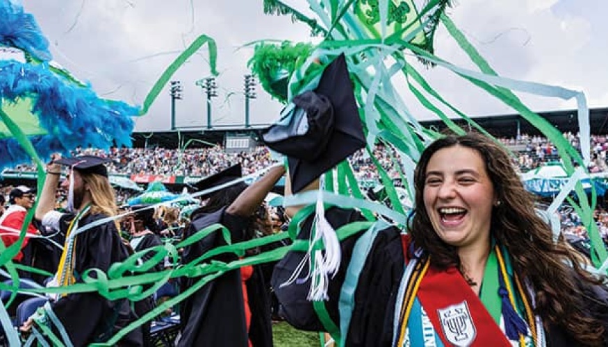 Crowd of graduates celebrate as streamers come down on a stadium field. A graduate in the foreground holds decorated umbrella.