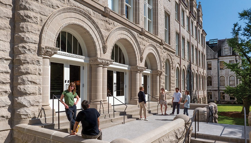 Several people gather outside a historic stone building on Tulane University's campus, chatting in small groups. The building features arched doorways, decorative stonework, and tall windows, with a bright blue sky and green lawn in the background.