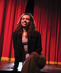 Lourdes Castillo sits on edge of stage with a red curtain in background