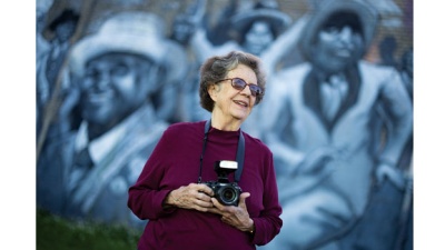 Judy Cooper holds her camera and stands in front of mural depicting second line dancers