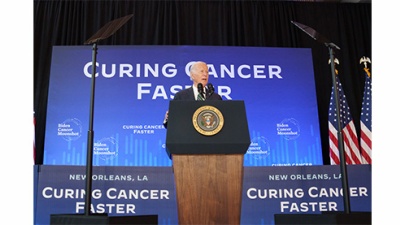 President Biden speaks from stage with signs that read Curing Cancer Faster 