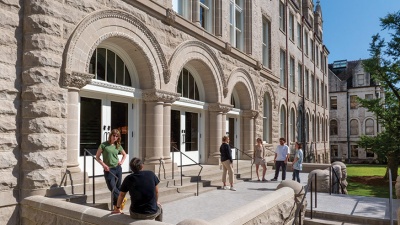 Richardson RMH Landing TH Several people gather outside a historic stone building on Tulane University's campus, chatting in small groups. The building features arched doorways, decorative stonework, and tall windows, with a bright blue sky and green lawn in the background.