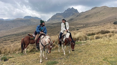 Altman Horses Two people ride horses on a grassy mountain landscape under a cloudy sky. They seem relaxed, engaged in conversation, with rugged peaks in the background.