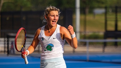 Student-Athletes Who Graduated with Honors A female tennis player wearing a white outfit passionately celebrates with a fist pump on an outdoor court, holding a red racket.