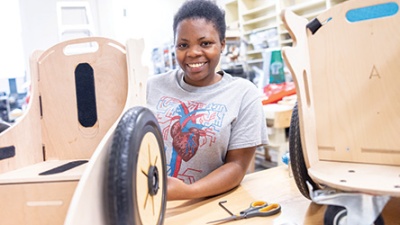 A student smiles while assembling a wooden mobility device at a workshop. The space is bright with tools and materials scattered around, conveying creativity.