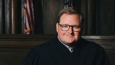 Cade R. Cole Justice Cade Cole in black judicial robes sits in a formal courtroom setting, wearing glasses and holding a book.