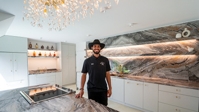 A smiling man in a cowboy hat stands in a modern kitchen with marble countertops and backsplash. Soft lighting creates a warm, inviting atmosphere.