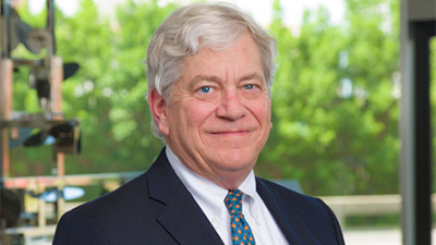 A man with white hair smiles warmly, wearing a dark suit with a blue polka dot tie in front of a window showing lush greenery outside.