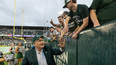 A man in a suit and cap high-fives enthusiastic fans at a football game. Cheerleaders stand nearby. The atmosphere is lively and celebratory.