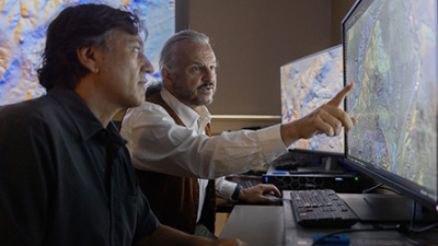 Two men analyze a detailed map on a large monitor. One points at the screen, focusing intently. The room is dimly lit, suggesting concentration.