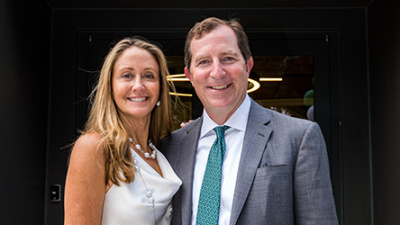 A smiling woman in a white dress and a man in a gray suit with a teal tie stand closely together, posing happily in front of a doorway.