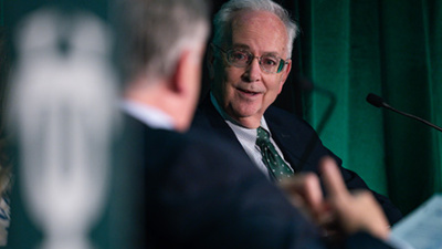 A man in glasses and a suit speaks at a panel, looking engaged, with a microphone nearby. A blurred person and green curtain are in the foreground.