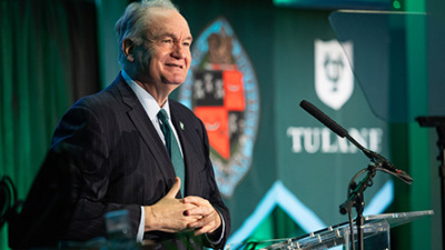 A man in a suit and tie speaks at a podium with a microphone. Behind him is a Tulane University banner. The atmosphere is professional and formal.