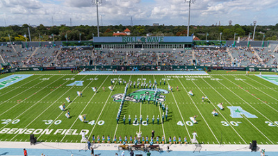Aerial view of a football stadium with a marching band forming a large letter "B" shape on the field. Spectators fill the stands, and the atmosphere is lively.