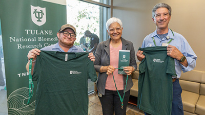 Three people stand smiling, holding green Tulane T-shirts. A Tulane National Biomedical Research Center banner is behind them, conveying a celebratory mood.