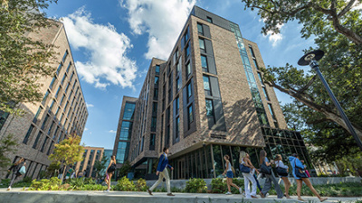 A group of students walks past modern, multi-story brick buildings under a blue sky with scattered clouds. The scene is lively and academic.