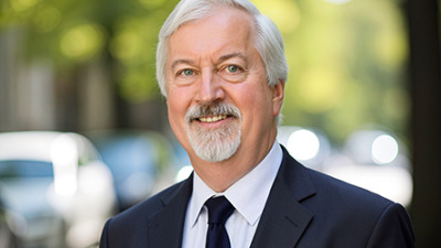 Bill Templeton in a suit smiling outdoors, with blurred trees and cars in the background. The scene conveys a professional and approachable tone.