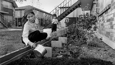 A woman tends to a garden with a watering can. A child stands nearby. Apartments with stairs are seen in the background.