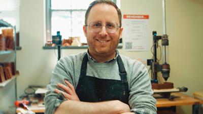 Smiling person with glasses and beard, arms crossed, wearing a black apron in a workshop. Shelves and tools create a cozy, industrious atmosphere.