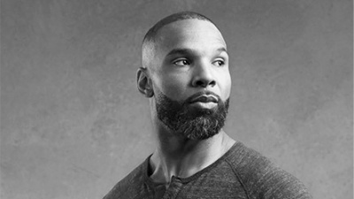 A black and white photo shows a man with a beard gazing to the side. He wears a dark, textured shirt, set against a plain background, conveying thoughtfulness.