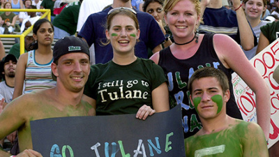Fans in a stadium cheer energetically, holding a sign that reads "Go Tulane Green Wave." Faces are painted green, expressing school spirit and excitement.
