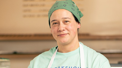 Smiling woman in a green bandana and "AYU BAKEHOUSE" apron, standing in a bakery.