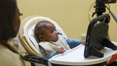 Baby in high chair watching a tablet, with an adult and camera nearby.