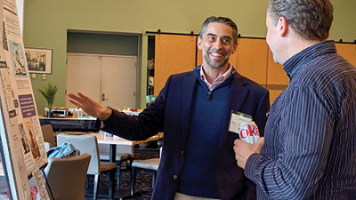 Smiling man in blue blazer explains poster to another man at an indoor event.