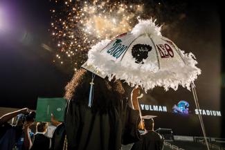 Fireworks light up the sky above Yulman Stadium as graduates watch. One graduate holds a decorated umbrella.