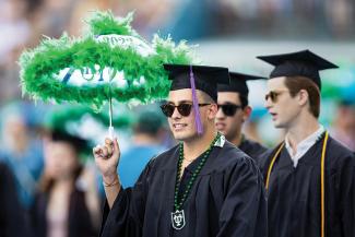 A graduate in cap and gown and sunglasses holds an umbrella decorated with green feathers.