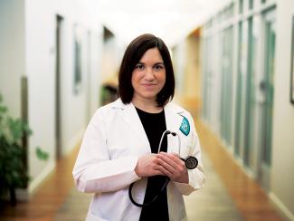  Dr. Kimberly Mukerjee stands in medical center hallway holding a stethoscope and wearing her white coat.