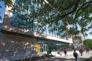 students walk on the sidewalk outside of the Paul Hall building, a tree casts a shadow 