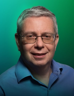 portrait of Lev Kaplan, wearing a blue shirt and glasses and standing against a green background